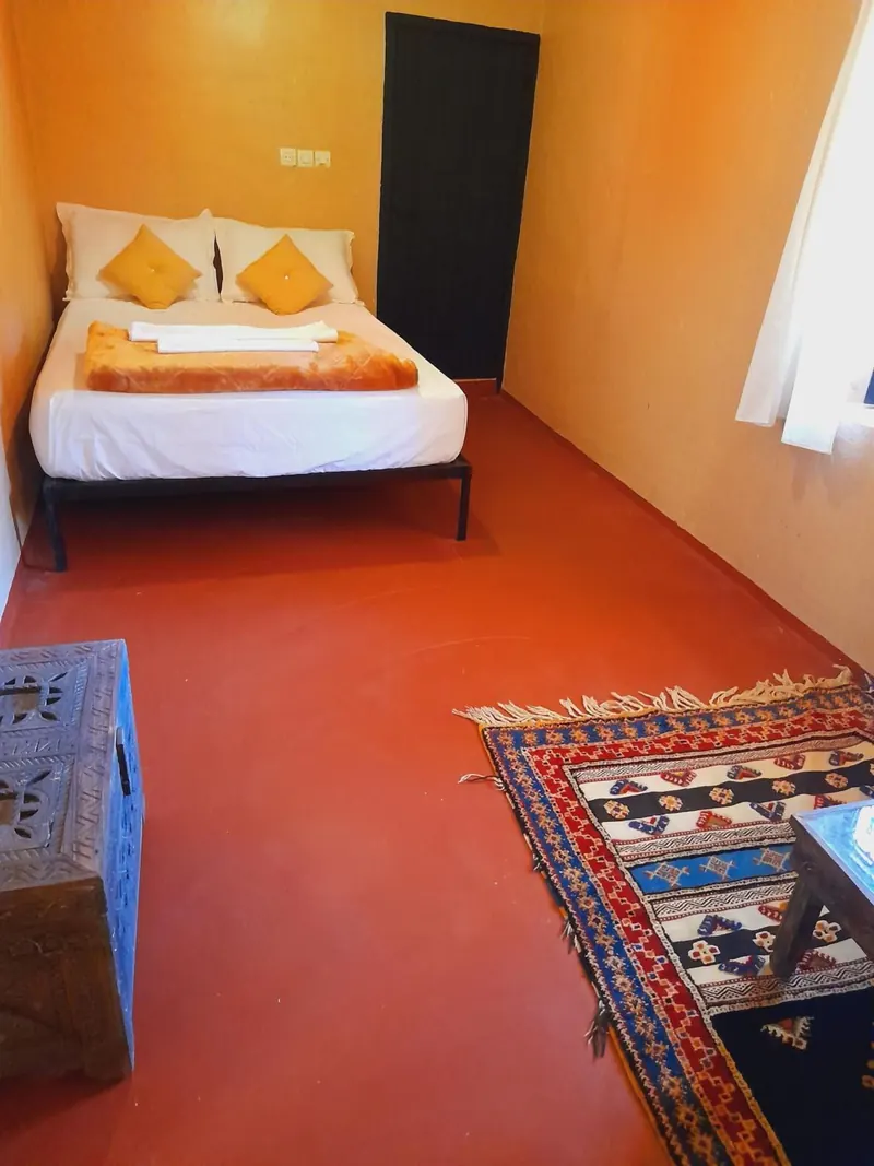 Double or Twin Room: a double bed with orange cushions, a carved chest and a rug